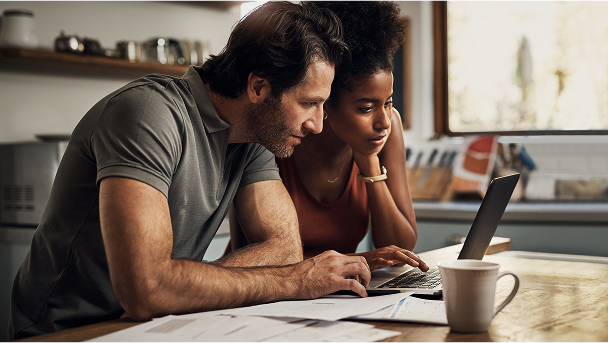A couple sitting in their kitchen looking at a laptop together with paperwork spread out on the table next to them.